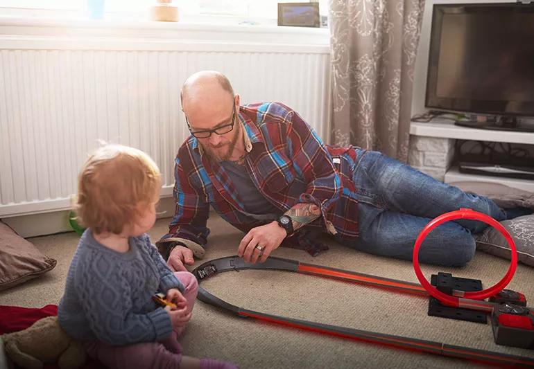 A balding man wearing a plaid shirt and a young child playing together with a toy car racetrack