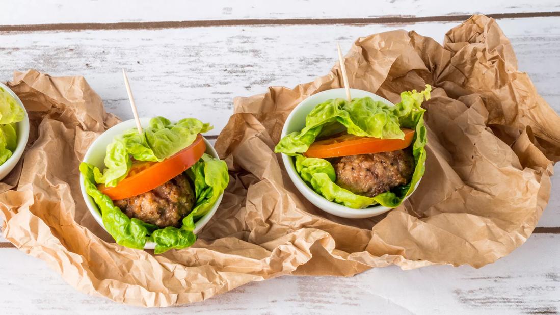 Lettuce-wrapped burgers with tomato, in white bowls atop crumpled brown bag