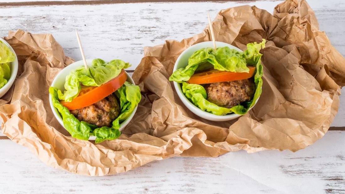 Lettuce-wrapped burgers with tomato, in white bowls atop crumpled brown bag