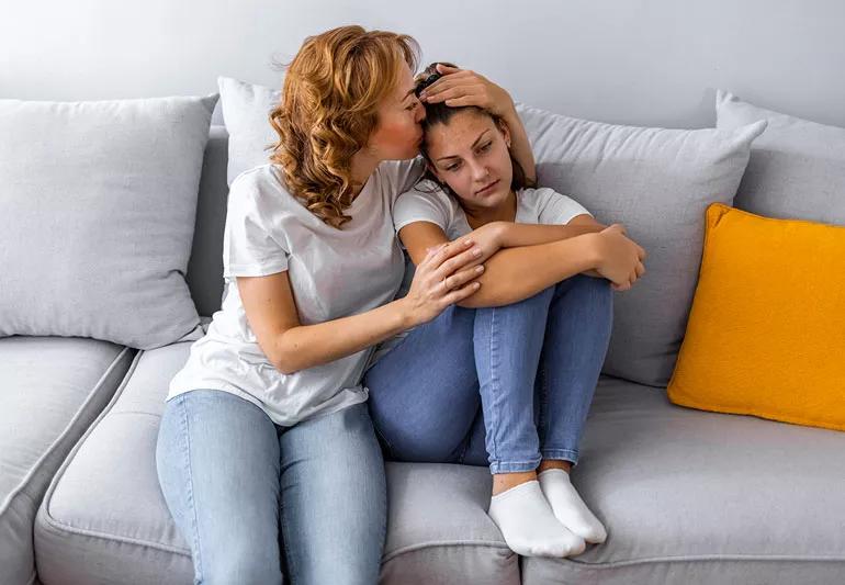 Parent comforts young teenager while both are sitting on couch.