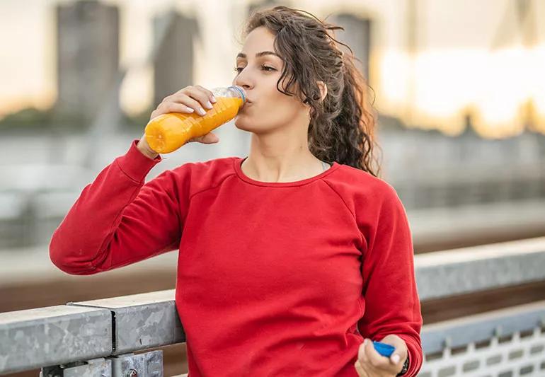 A woman wearing a red sweatshirt and drinking from a bottle of orange juice