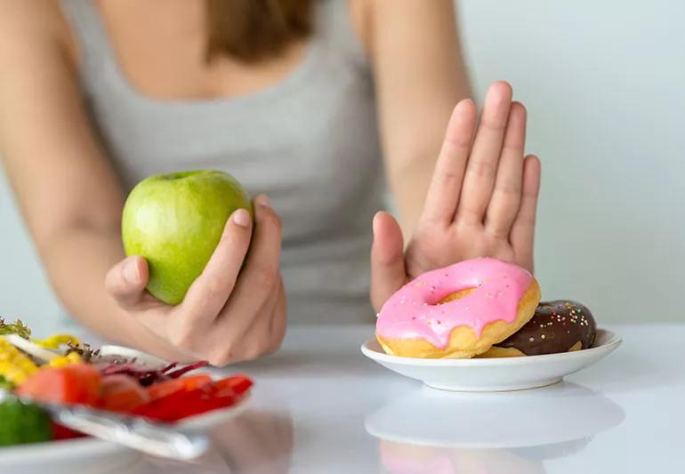 One hand holding an apple while another pushes away a plate full of iced donuts