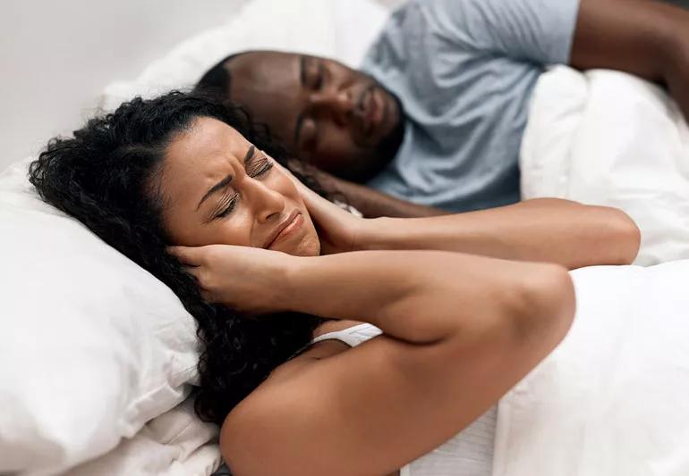 woman covering ears while husband snores in bed