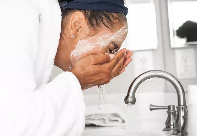 Person washing cleasning product off of their face over wash basin in white bathroom.