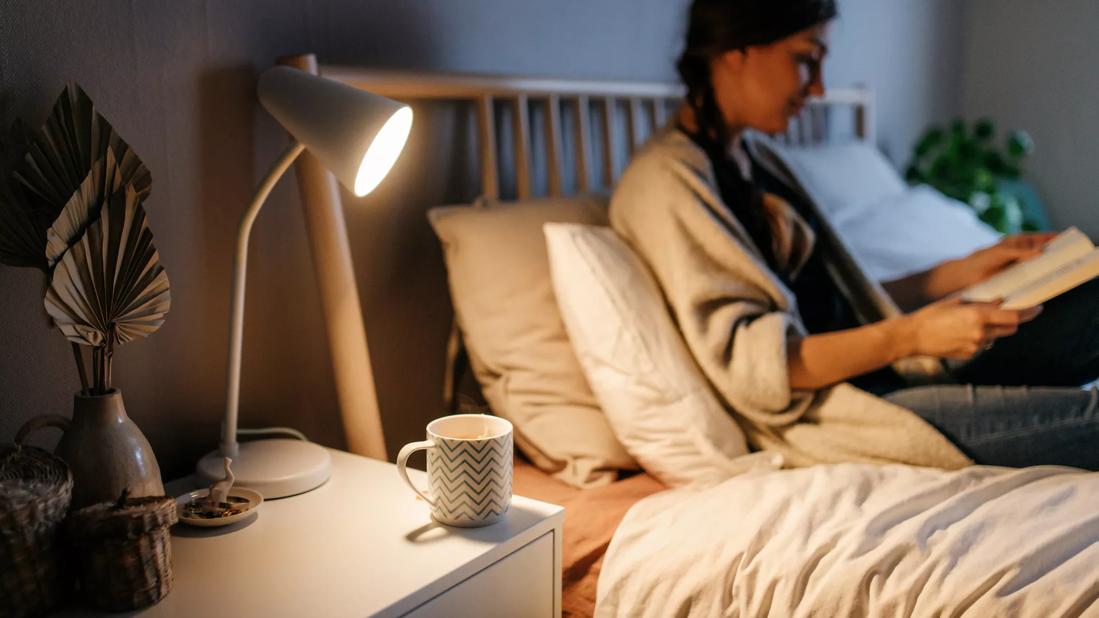 Person sitting in bed in the evening, reading a book, with cup of tea on bedside table