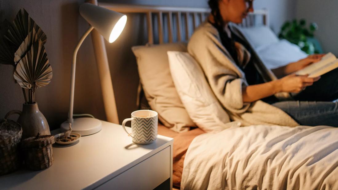 Person sitting in bed in the evening, reading a book, with cup of tea on bedside table