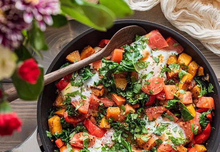 A pan of butternut squash mixed with tomatoes, eggs and green herbs