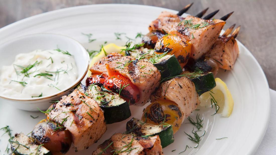 Salmon and vegetable kebabs covered in herbs next to a small dish of white sauce