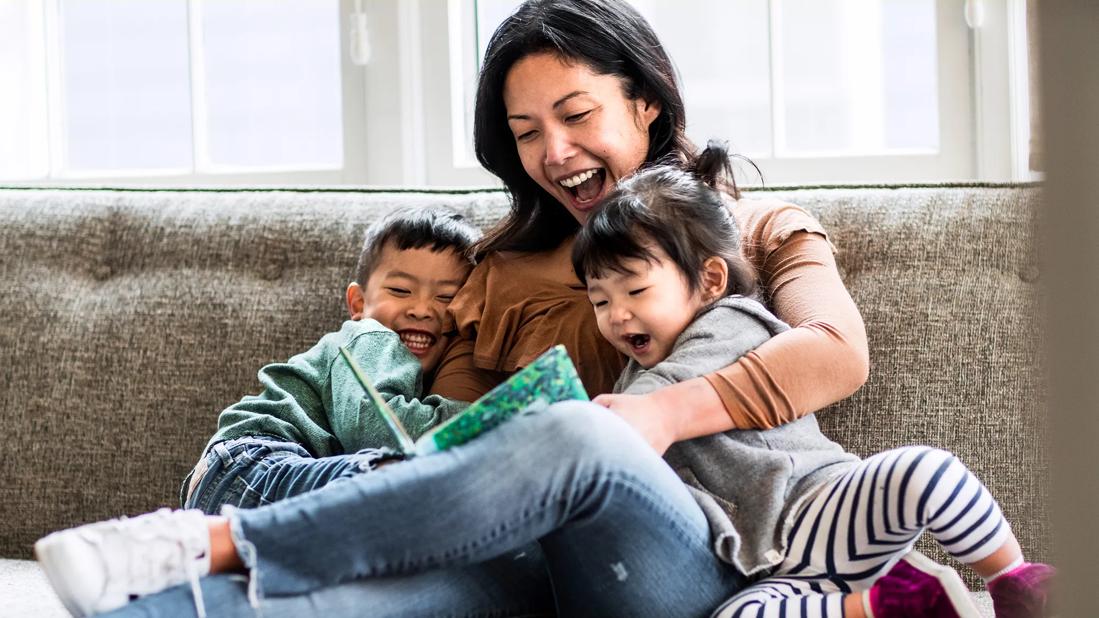 Caregiver laughing and reading to two happy kids on couch