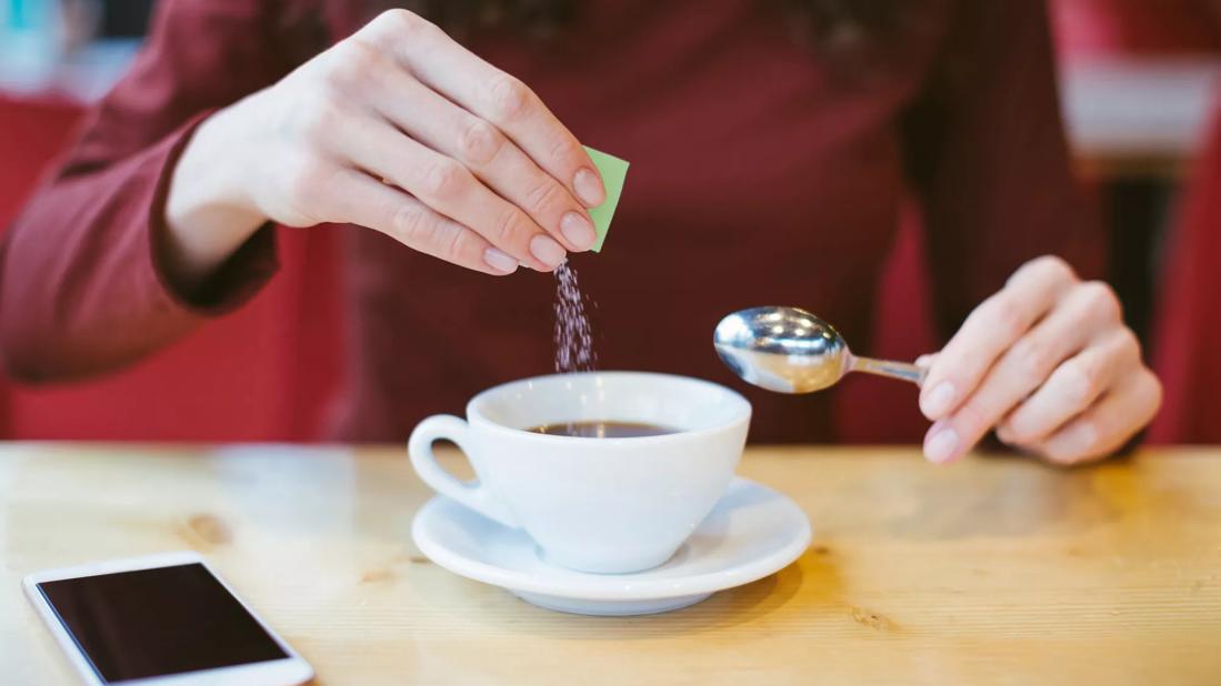 Person pouring packet of sugar subsitute in cup of coffee, cell phone on table, spoon in hand
