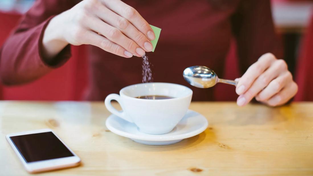 Person pouring packet of sugar subsitute in cup of coffee, cell phone on table, spoon in hand