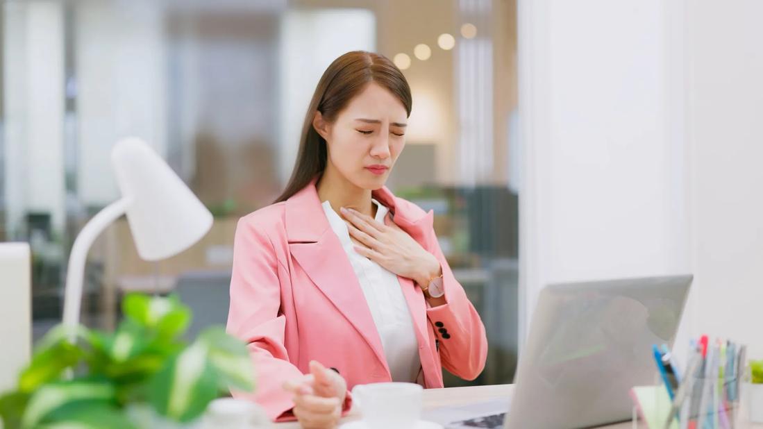 Person at desk at work, eyes closed in discomfort, hand at base of throat