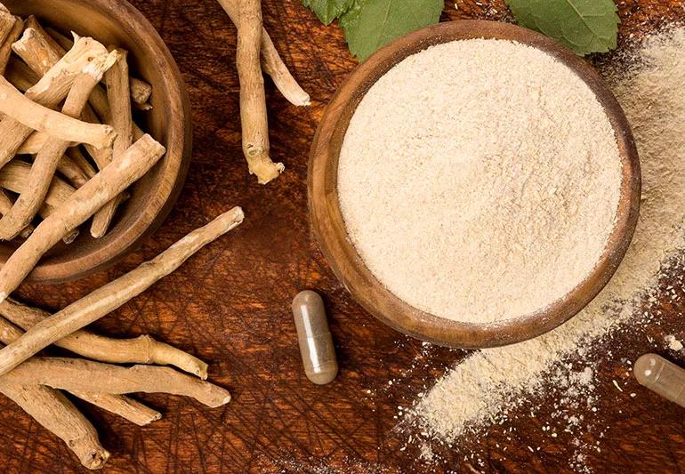 A bowl of cream-colored Ashwagandha powder, a brown root and olive-colored capsules