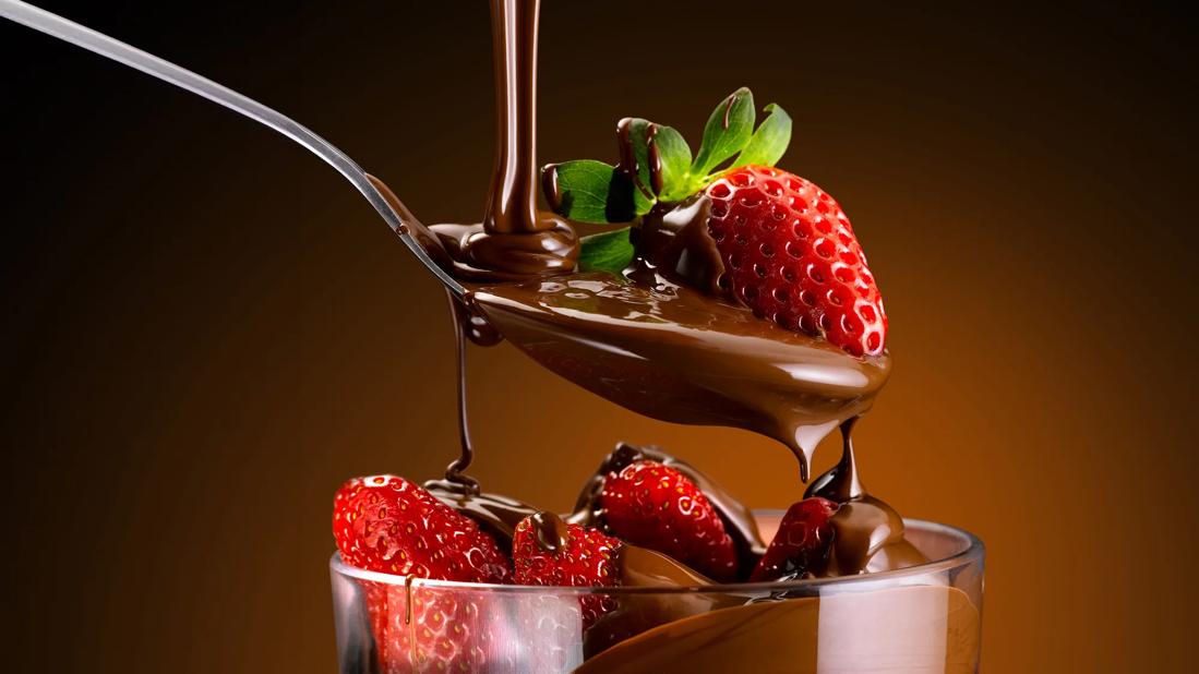 Spoon with a strawberry, with chocolate drizzling down over it, and drizzling down over more strawberries in a glass dish