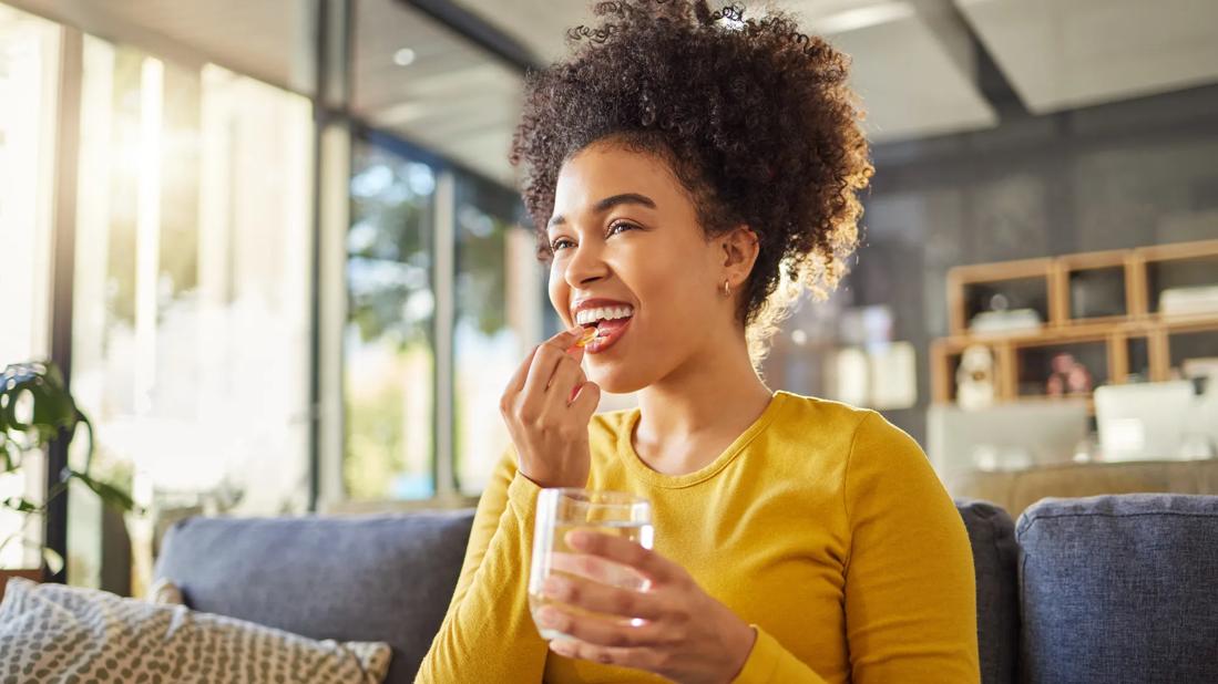 Young person smiling and holding a pill to their mouth while holding a glass of water in their other hand