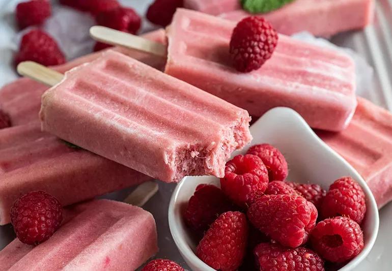 Red frozen popsicles are stacked next to a small white bowl of red raspberries.