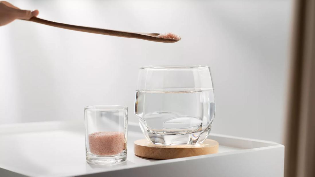 Wooden spoon with pink Himalayan salt over glass of water, with container of pink Himalayan salt