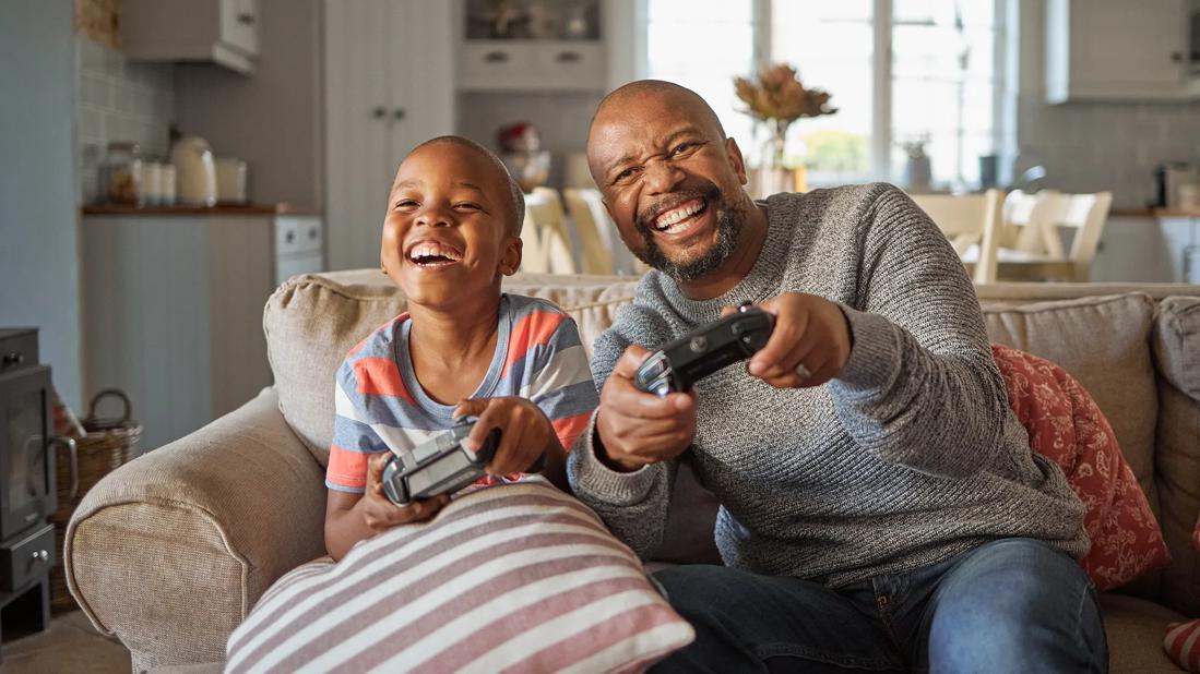 Happy, laughing child and caregiver sitting on couch holding video game controllers, playing video games