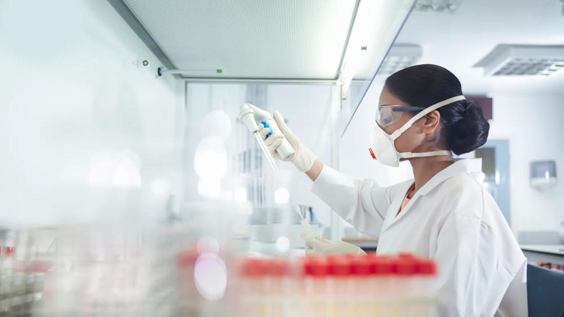 Scientist/lab tech in lab wearing mask and white coat working, with test tubes on counter