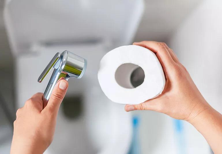 A person holds up a bidet in their left hand and a roll of toilet paper in their right hand.