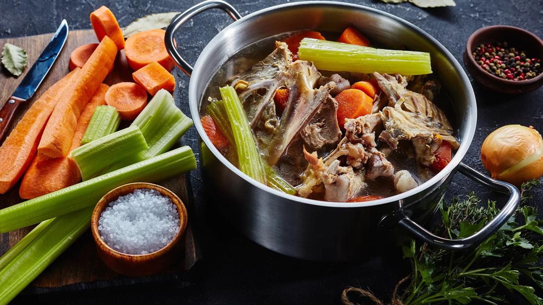 Steel pot of bone broth mixed with carrots and celery, with carrots and celery on cutting board and seasonings