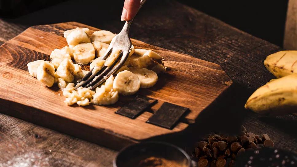 Mashing bananas with fork on wooden cutting board, with chocolate chips on table