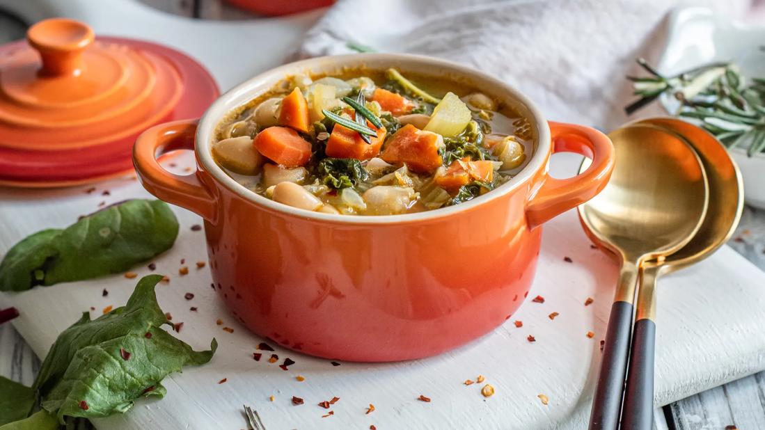 A bright orange ceramic pot full of white bean stew with rosemary and spinach, with spoons and crock lid on table