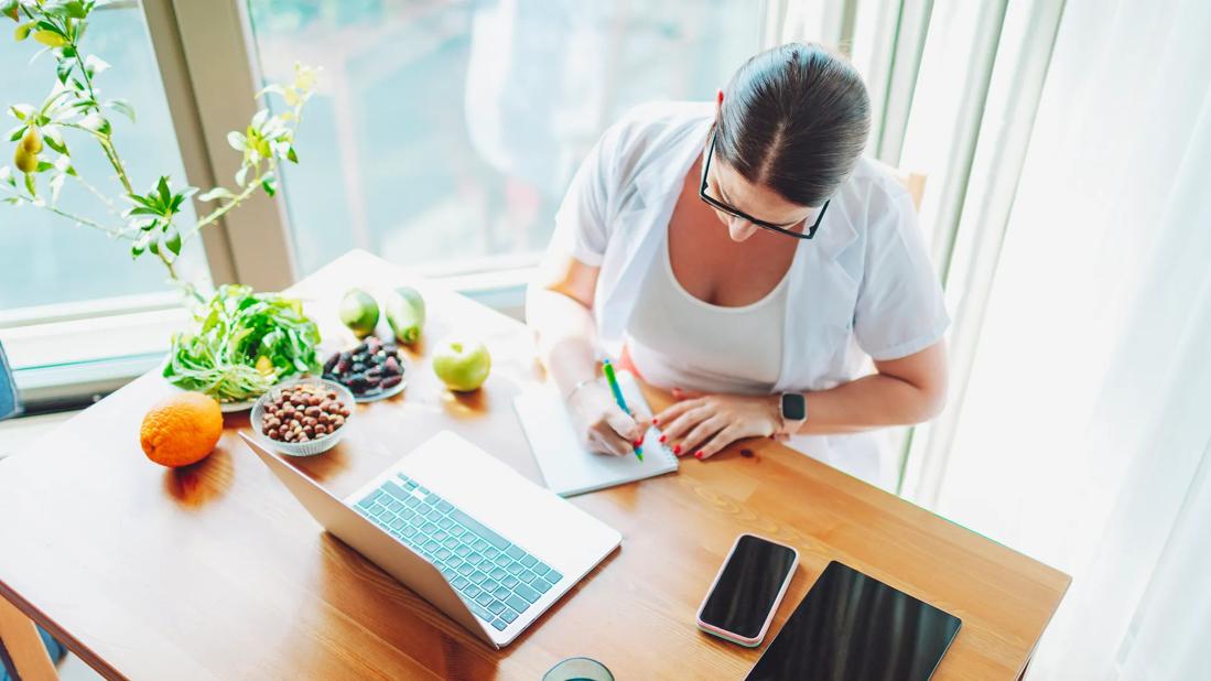 Person journaling at desk in sunny home office, with laptop, smartphone and healthy fruits and nuts on desk