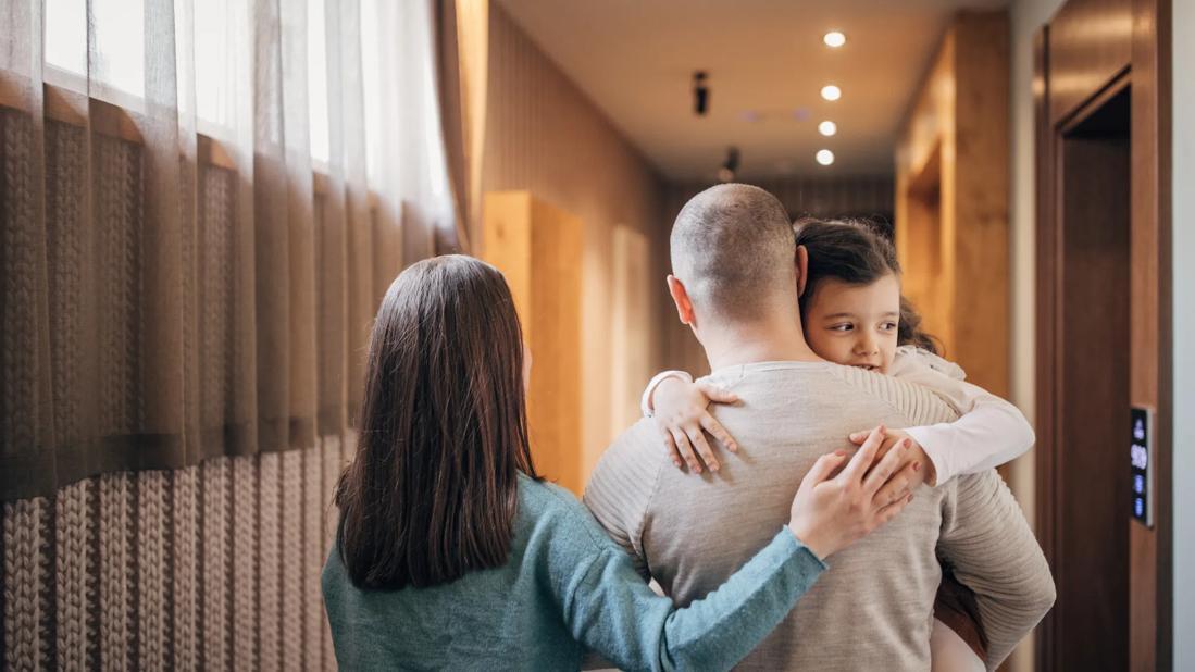 Caregiver carrying child, with other caregiver walking with arms around them, walking down hotel hallway