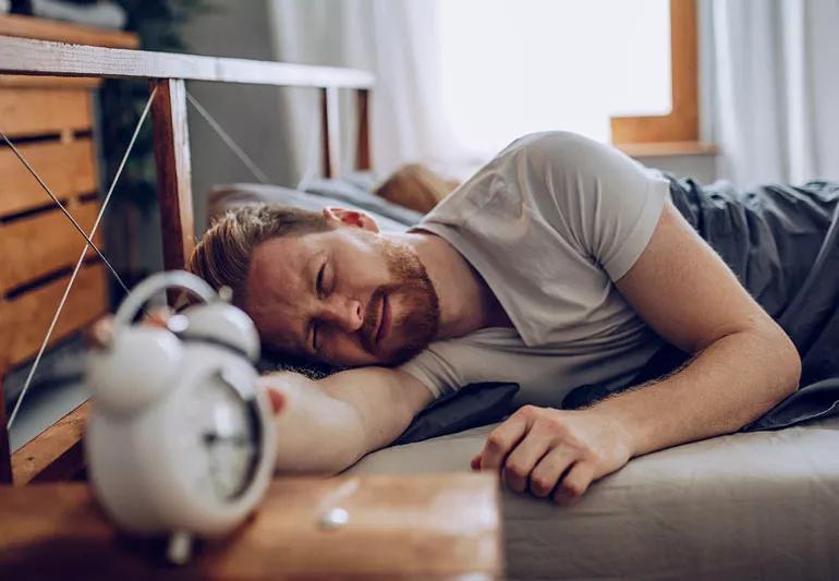 Man hitting snooze button on his alarm clock while in bed