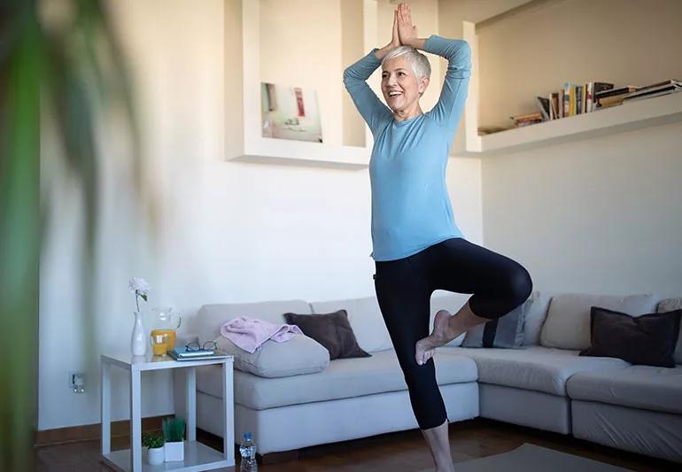 Senior balancing on one foot with hands above head in a living room