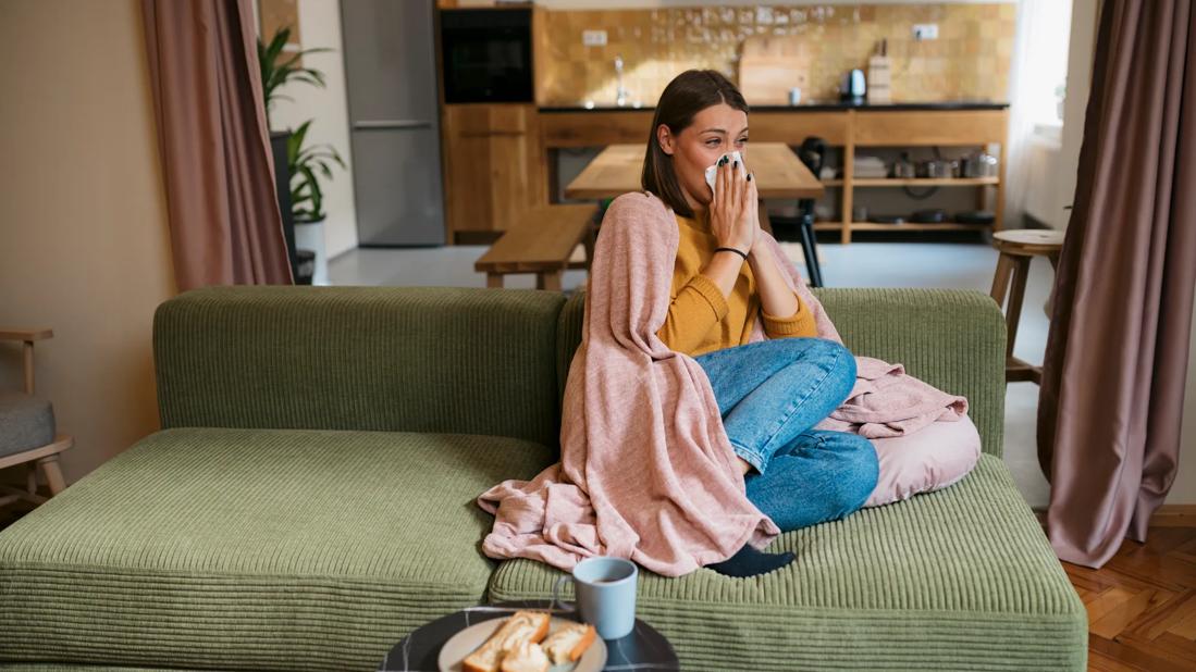 Person sitting on couch at home, wrapped in blanket, blowing their nose, with toast and tea on table