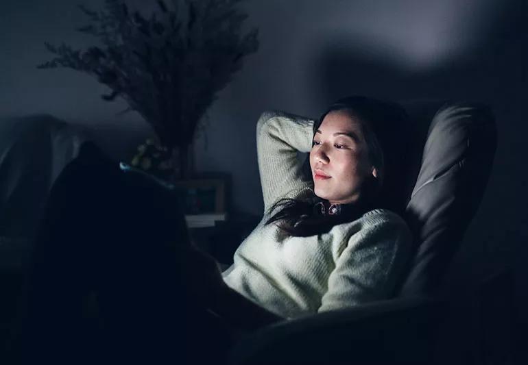 A woman relaxing in a dark room who is lit only by the glow of a computer tablet