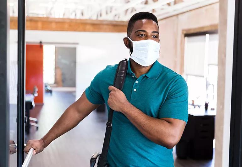 A man wearing a white surgical mask opening a door and holding a work bag over his right shoulder