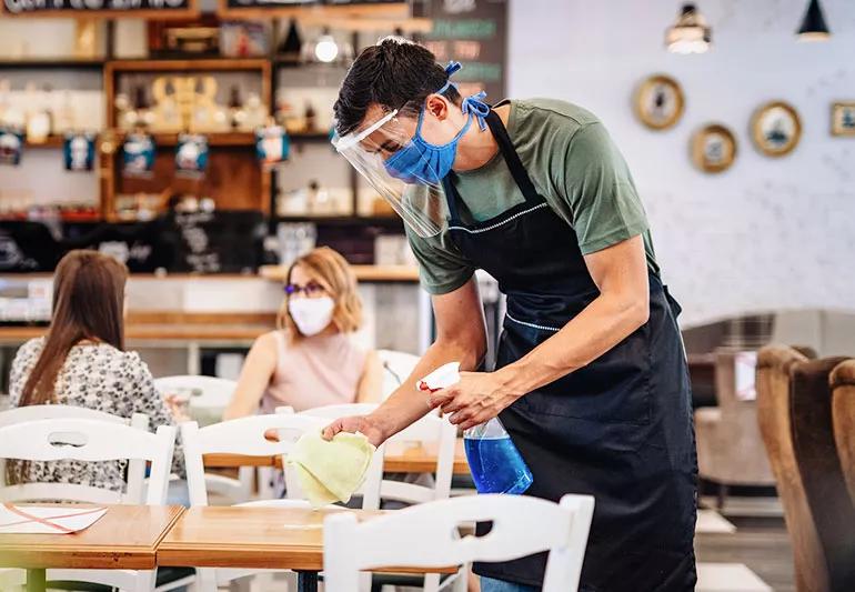 server wiping down tables in restaurant while wearing mask and face shield