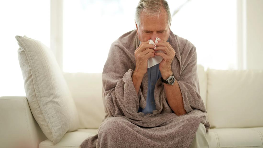 Older person sitting on couch, wrapped in blanket, hunched over, blowing their nose