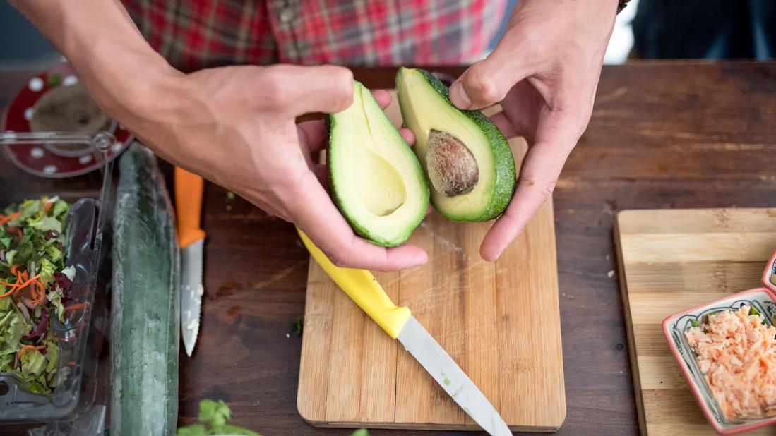 Person holding sliced open avocado halves over cutting board, among other food prep