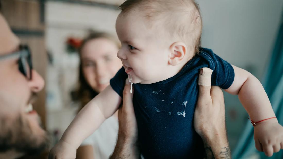 Smiling caregiver holding up smiling baby with spit-up on face and onsie, with smiling caregiver in background, too