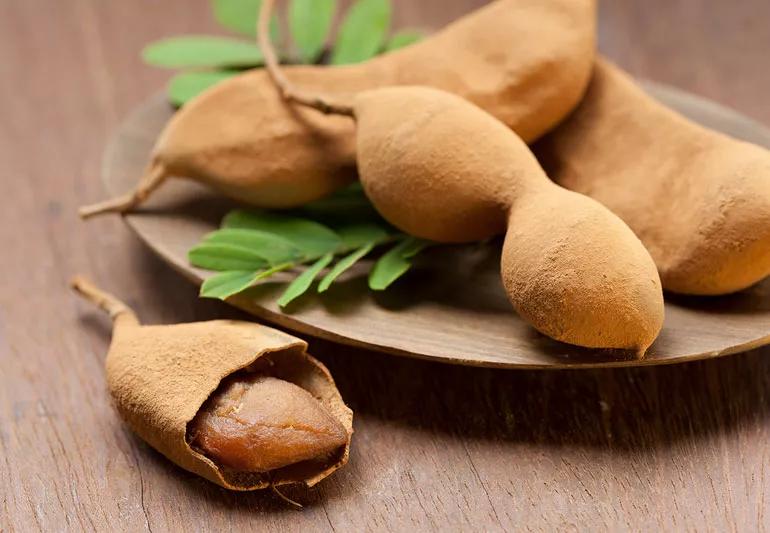 Tamarind pods displayed on brown plate on a wooden table, with one pod torn open showing fruit inside.