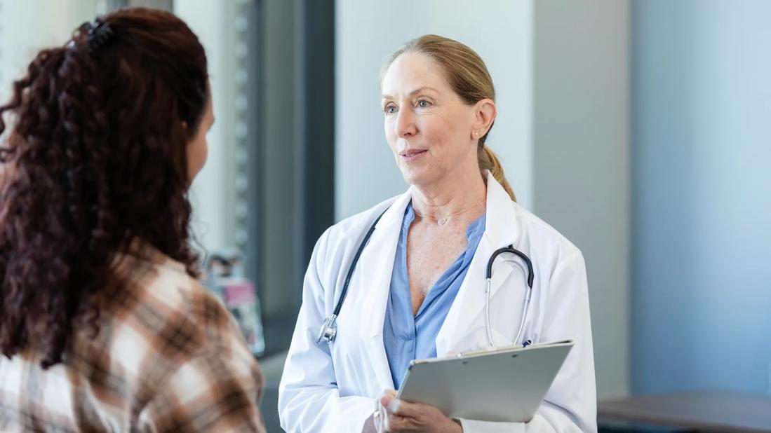 Healthcare provider, with stethoscope around neck, holding clipboard, talking to person in their office