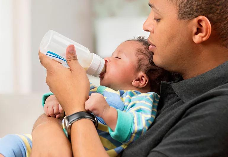 Parent feeds sleepy baby a bottle, while holding them in thier arms.