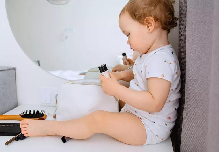 Curious toddler exploring mom's makeup case, holding foundation tube.