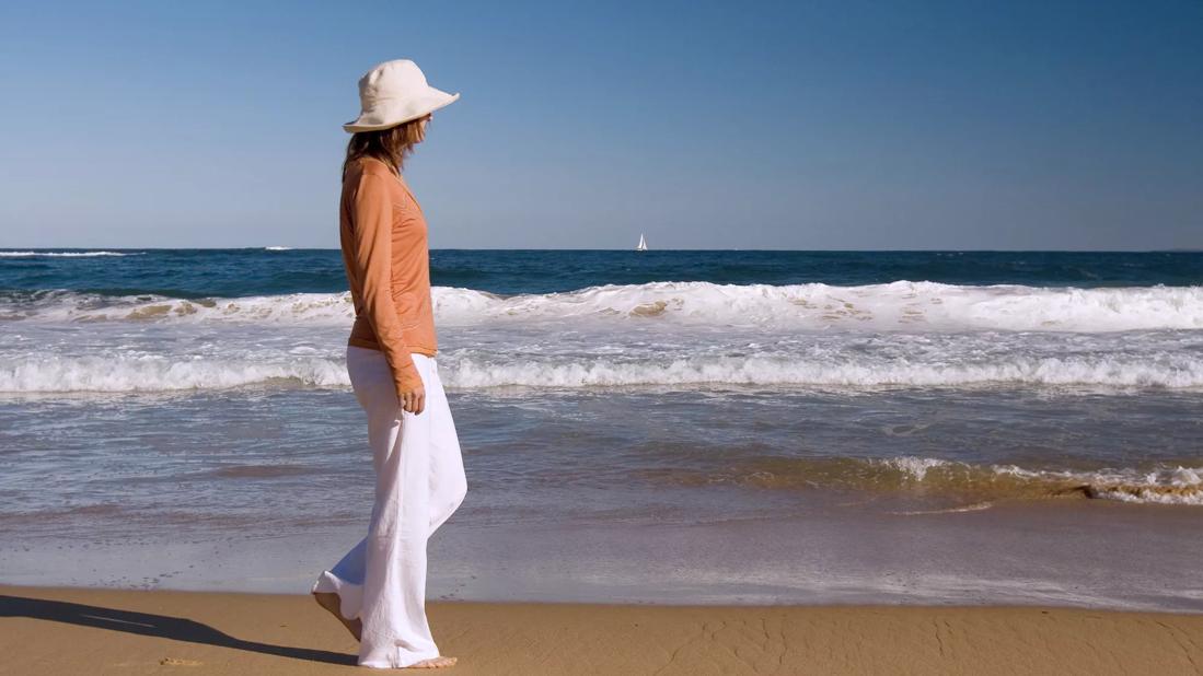 person walking along shore wearing sunhat and long-sleeve clothing