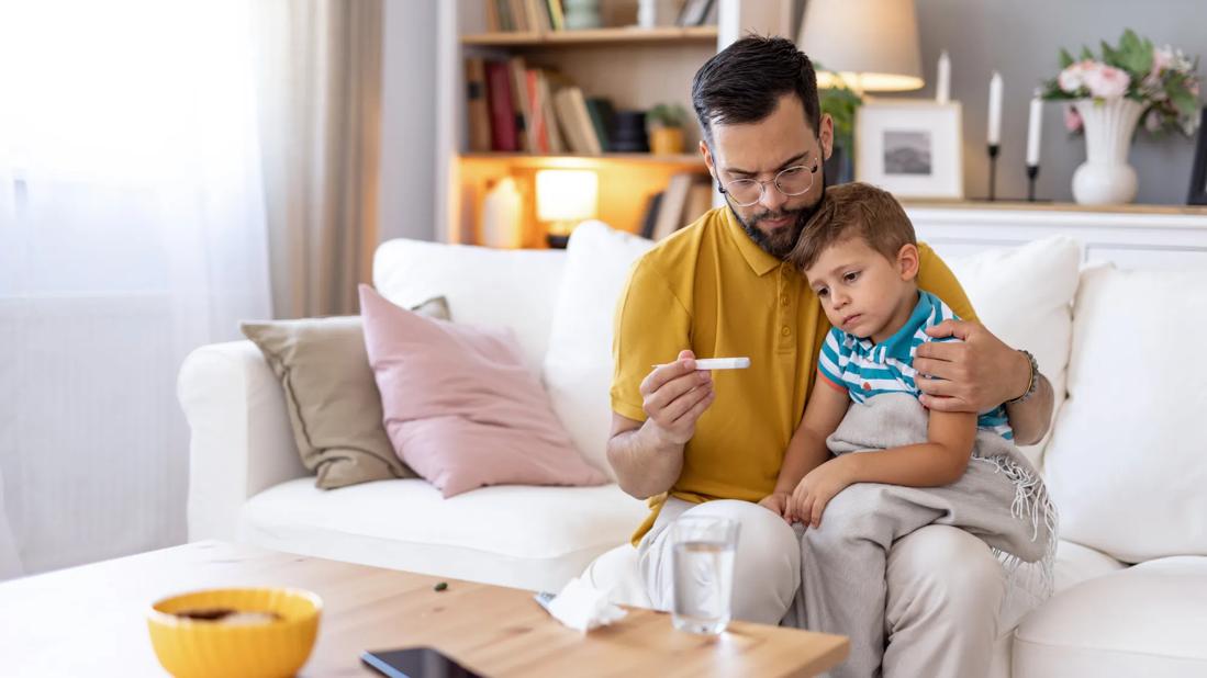 A parent checks a child’s thermometer while sitting on a couch