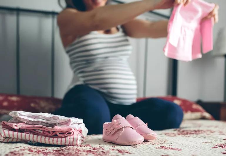 Pregnant woman sitting on bed sorting through baby clothes