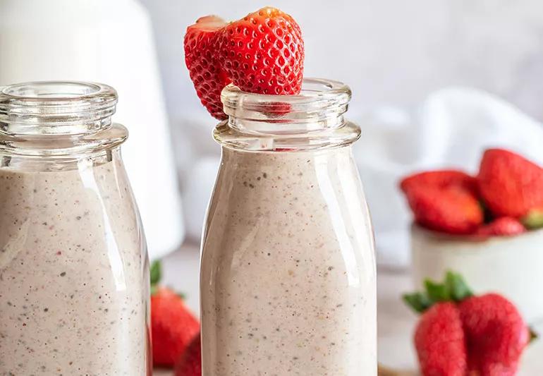 Cream-colored smoothies in thin glass bottles with strawberries in background and on rim