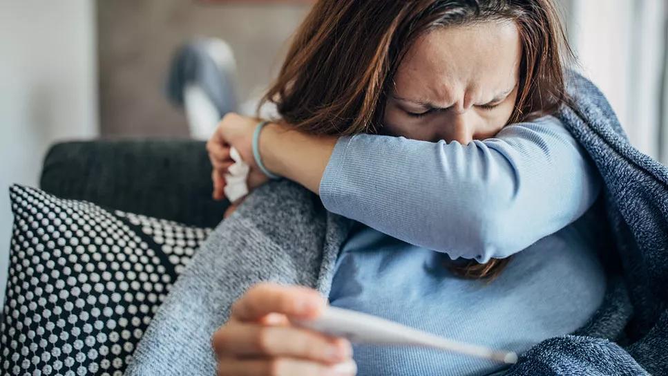 Adult female on couch, coughing into crook of arm, holding thermometer