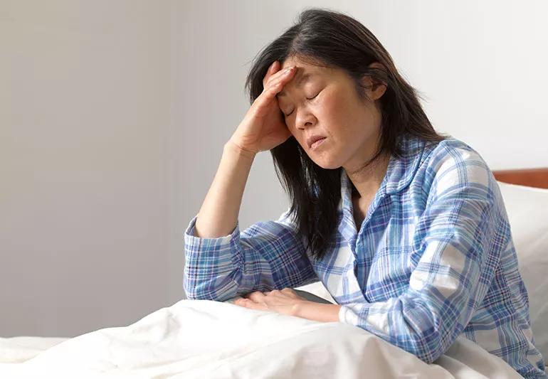 A woman wearing pajamas sitting in bed with her eyes closed, holding her head like she's in pain