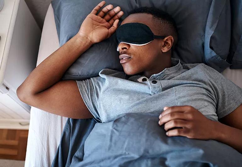 Person asleep on their back in bed with an eye mask on and one arm above their head