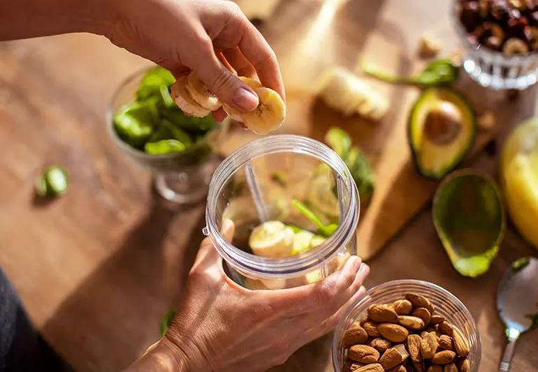 Person dropping sliced bananas into a cup to make a smoothie, with other foods in the background